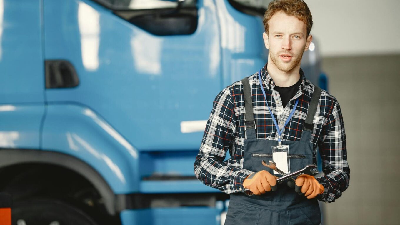 Young auto mechanic holding a wrench in an automotive workshop with a truck in the background.